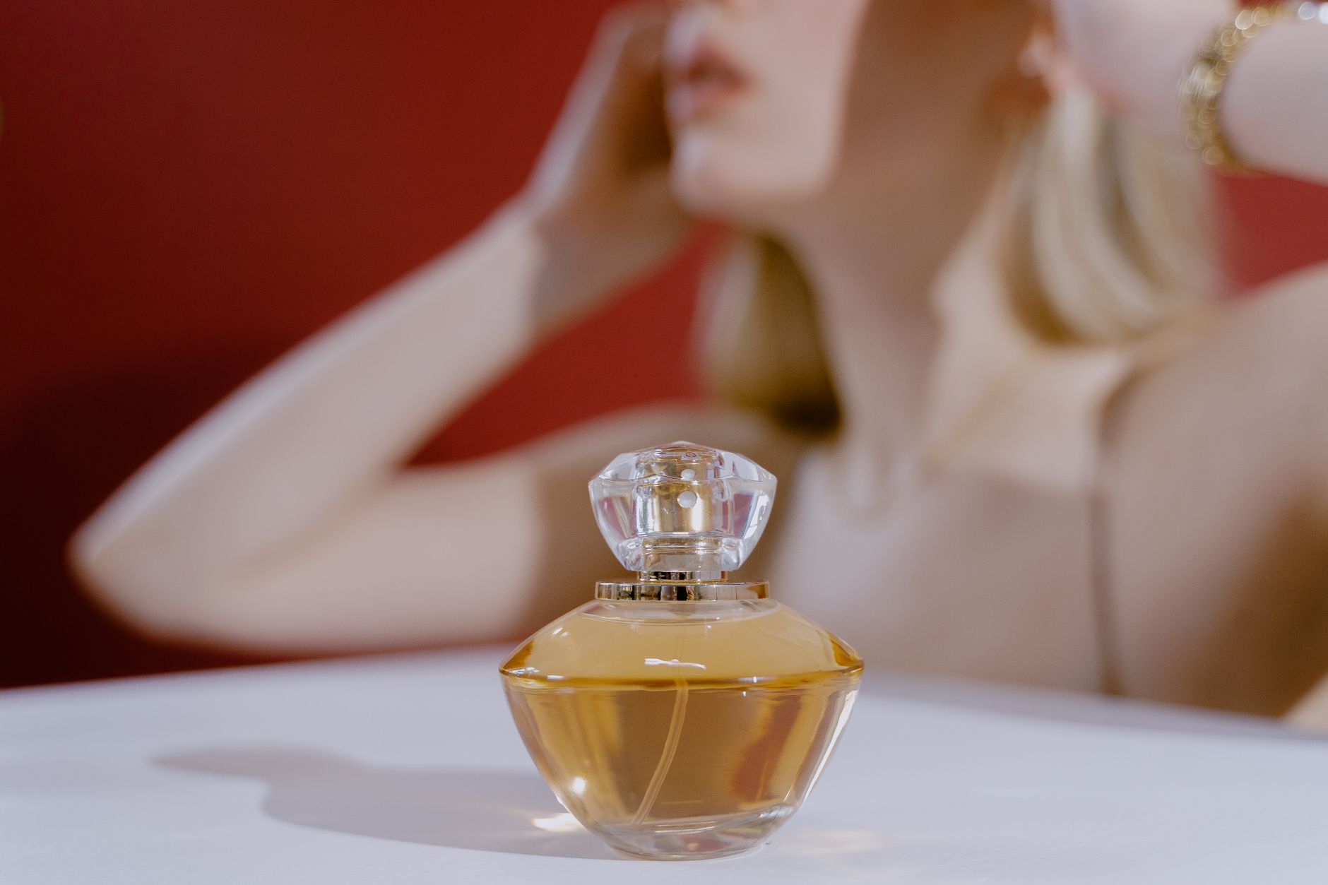 Przezroczysta butelka z żółtawym perfumem i krystaliczną korką na białym tle, rozmazana postać kobiety w tle