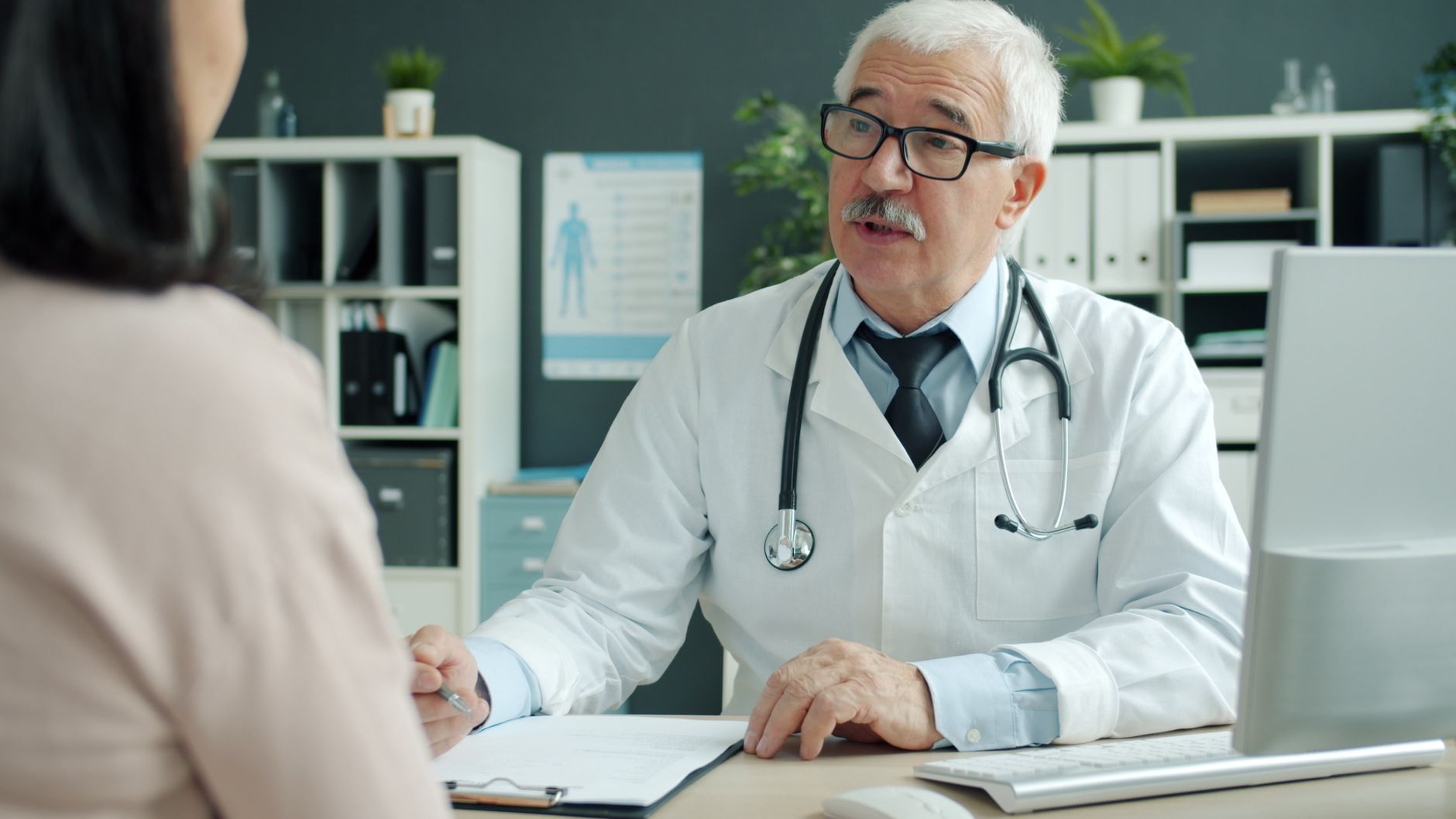 Starszy lekarz w białym kitlu i okularach rozmawia z pacjentem przy biurku w gabinecie