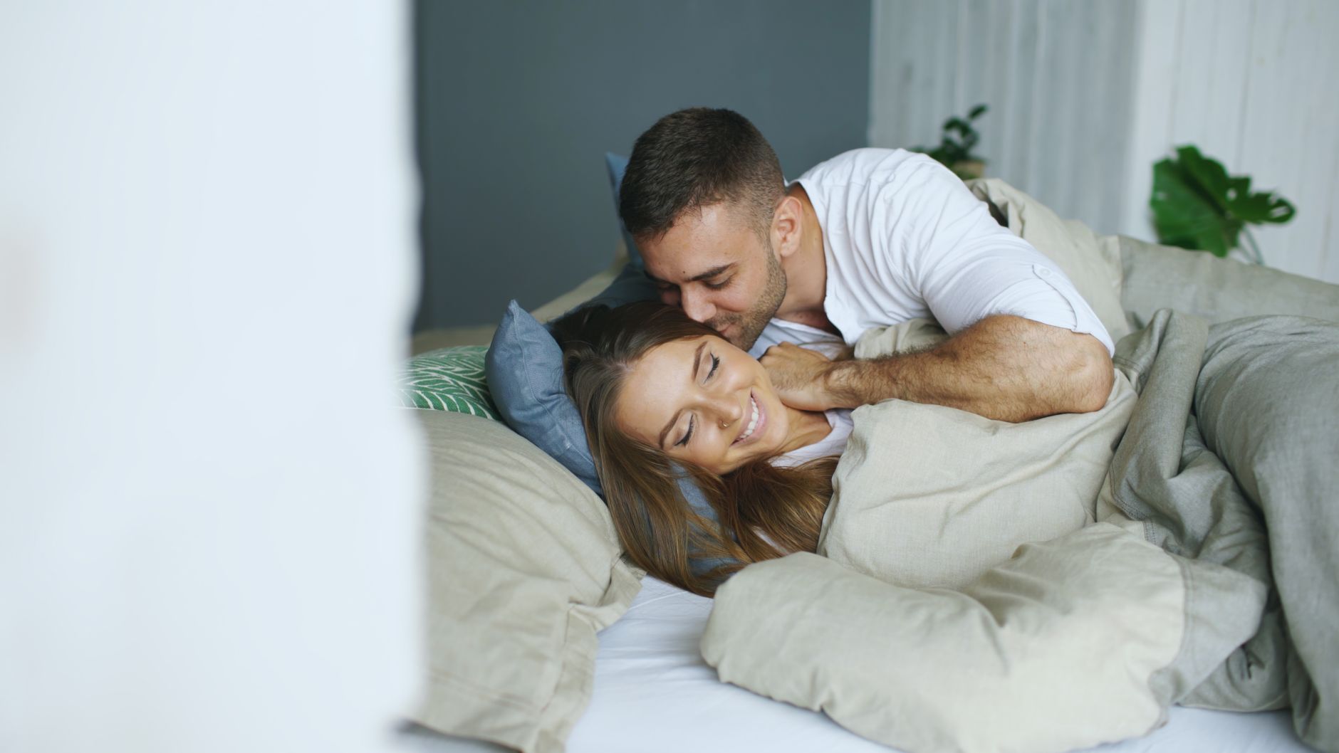 Młoda para przytulająca się na łóżku w jasnym, przytulnym wnętrzu z zieloną rośliną