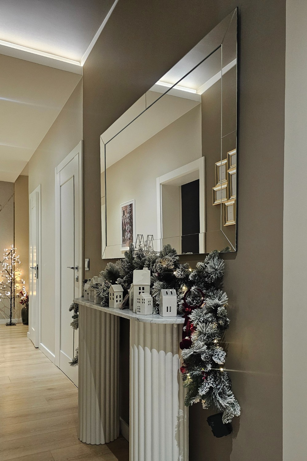 Eleganckie lustro prostokątne z białą metalową ramą nad białym grzejnikiem ozdobiony świąteczną girlandą i białymi ceramicznymi domkami